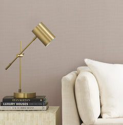 A gold desk lamp and stacked books sit on a light side table next to a cream-colored sofa with a white pillow, set against a wall adorned with Gossamer Woven Wallpaper by York Wallcoverings.