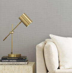 A gold desk lamp sits on a small table next to a beige sofa with a white pillow, with several books stacked under the lamp. The background features York Wallcoverings’ Gossamer Woven Wallpaper in a neutral hue, adding subtle texture to the space.