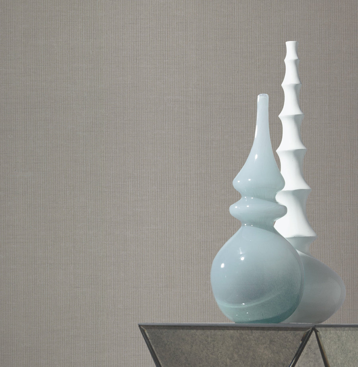 Two decorative vases, one light blue with a glossy finish and curved shape, and one white with a tiered, sculptural design, sit on a glass table against a plain backdrop featuring York Wallcoverings’ Gossamer Woven Wallpaper.