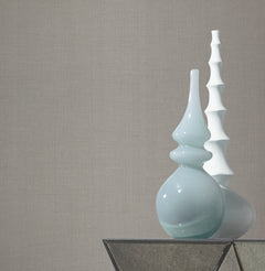 Two decorative vases, one light blue with a glossy finish and curved shape, and one white with a tiered, sculptural design, sit on a glass table against a plain backdrop featuring York Wallcoverings’ Gossamer Woven Wallpaper.