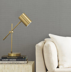 A modern gold desk lamp on stacked books beside a beige sofa with a white pillow, set against a backdrop of Gossamer Woven Wallpaper by York Wallcoverings.