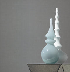 Two modern ceramic vases—one glossy light blue with smooth curves and one matte white featuring a ridged, geometric design—stand side by side on a glass table against a gray Gossamer Woven Wallpaper by York Wallcoverings.
