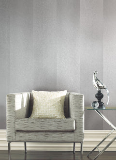 A modern armchair with a textured pillow sits beside a glass side table topped with a metallic bird sculpture, all set against York Wallcoverings’ Etched Chevron Wallpaper and dark wood flooring.