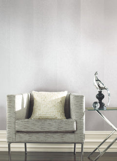 A modern gray armchair with a white textured pillow sits beside a glass side table displaying a decorative glass bird sculpture, set against York Wallcoverings’ Etched Chevron Wallpaper.