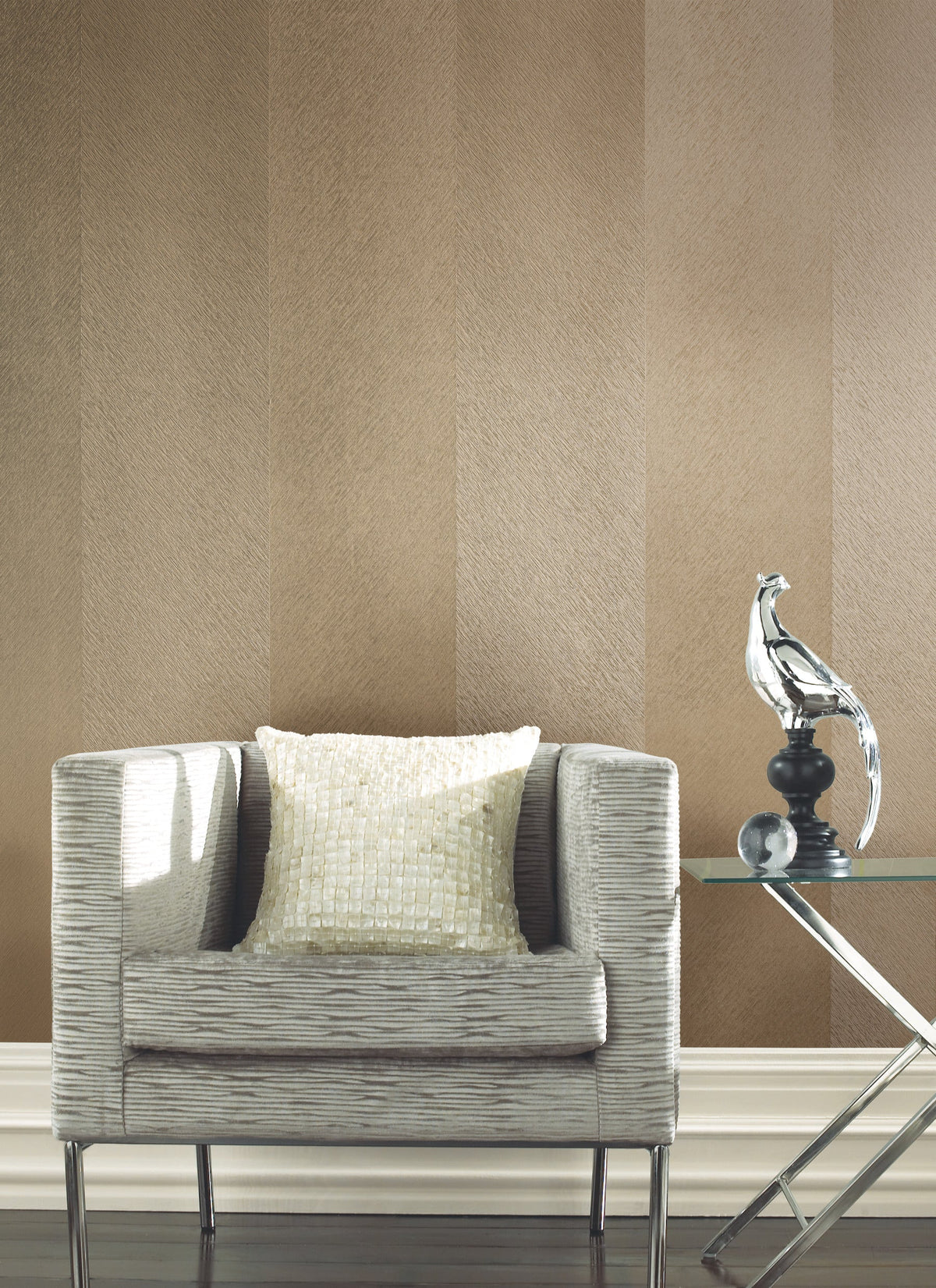 Set against a feature wall covered in York Wallcoverings’ Etched Chevron Wallpaper, a modern gray armchair with a textured cushion sits beside a glass table topped with a silver bird sculpture.
