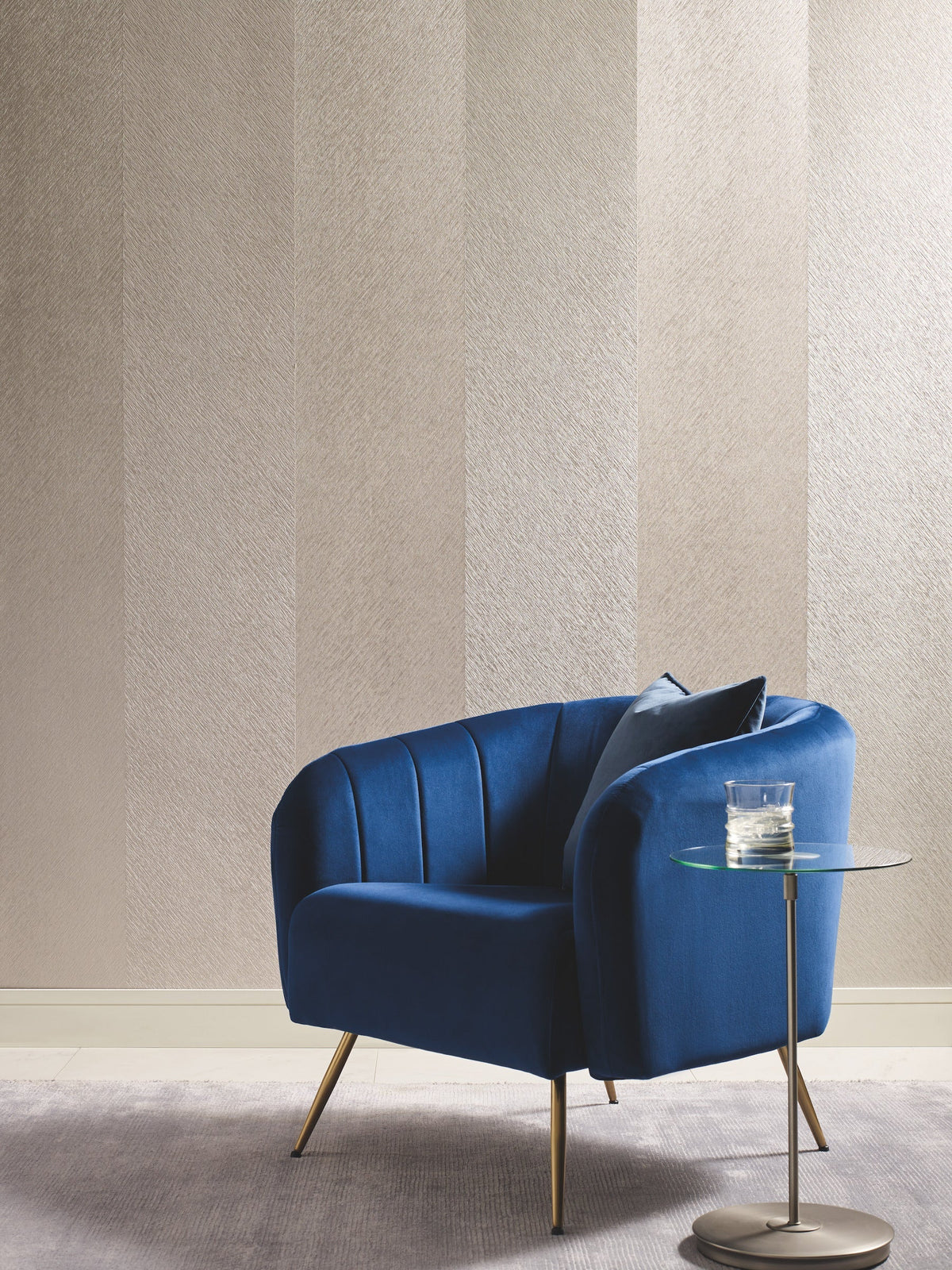 A modern room with a blue armchair, a small round glass side table holding a glass of water, and York Wallcoverings’ Etched Chevron Wallpaper featuring subtle texture and stripes in the background. A gray rug covers the floor.