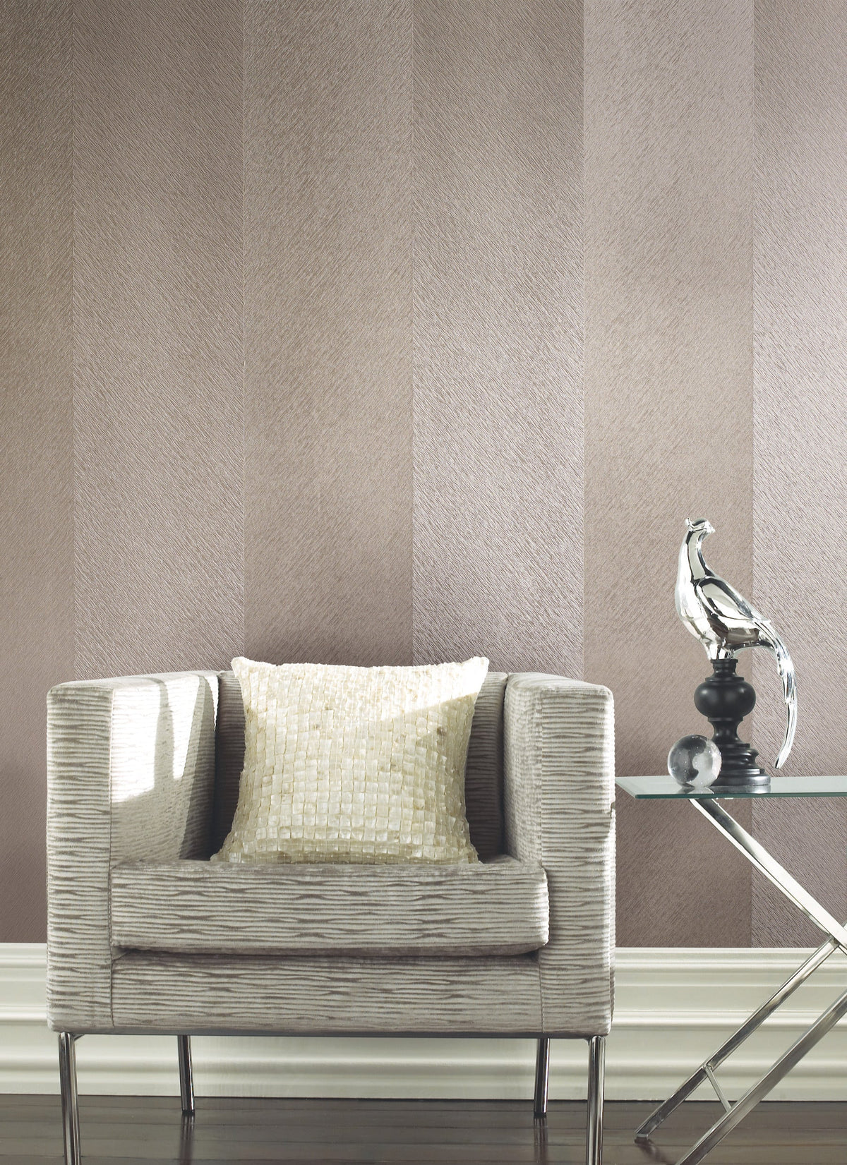 A textured beige armchair with a square sequined pillow sits beside a glass side table displaying a metallic bird sculpture, set against York Wallcoverings’ Etched Chevron Wallpaper.