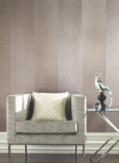 A textured beige armchair with a square sequined pillow sits beside a glass side table displaying a metallic bird sculpture, set against York Wallcoverings’ Etched Chevron Wallpaper.