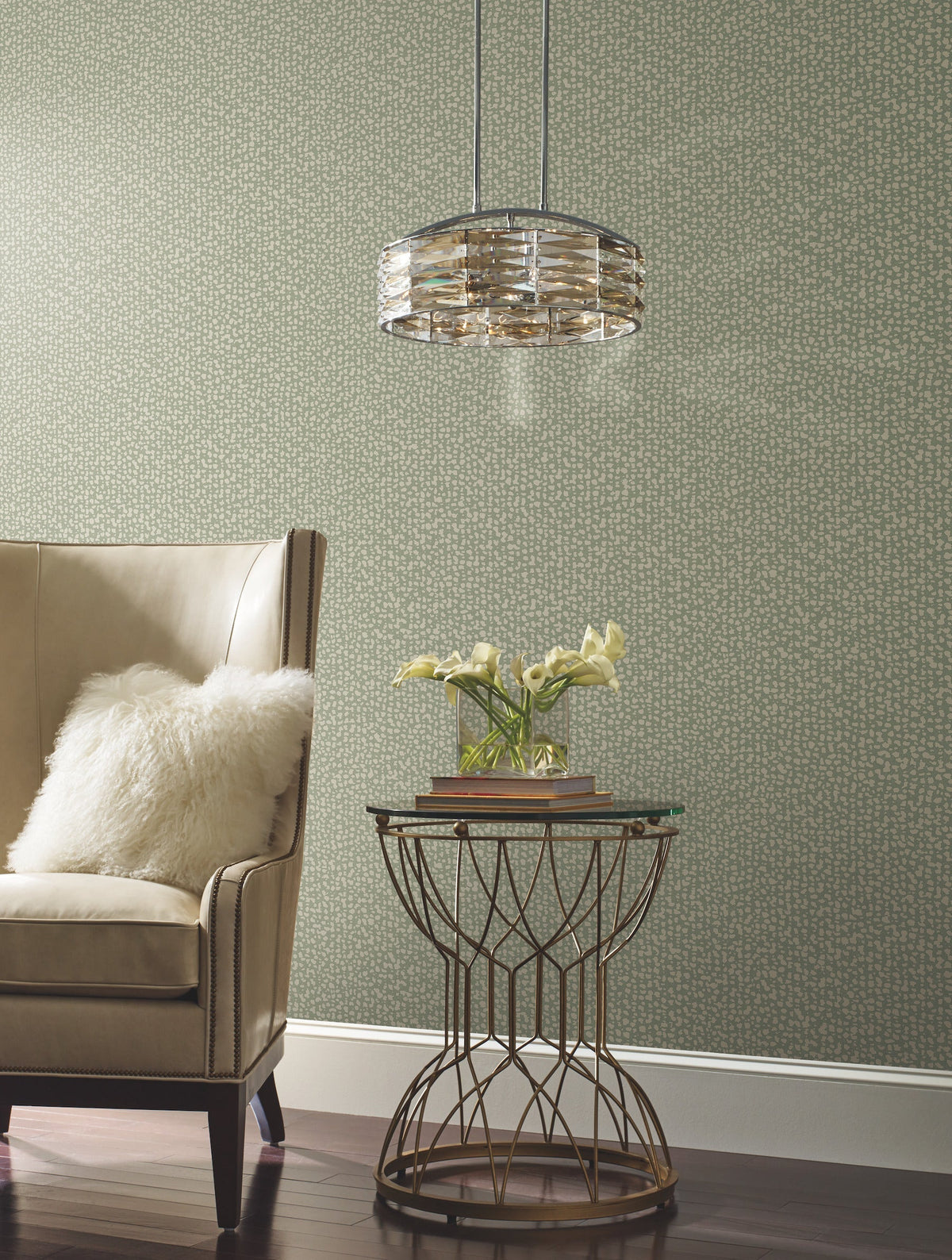 A beige armchair with a fluffy white pillow sits next to a round gold side table holding a small vase of white flowers and books, beneath a modern hanging light, set against Galaxies Wallpaper by York Wallcoverings.