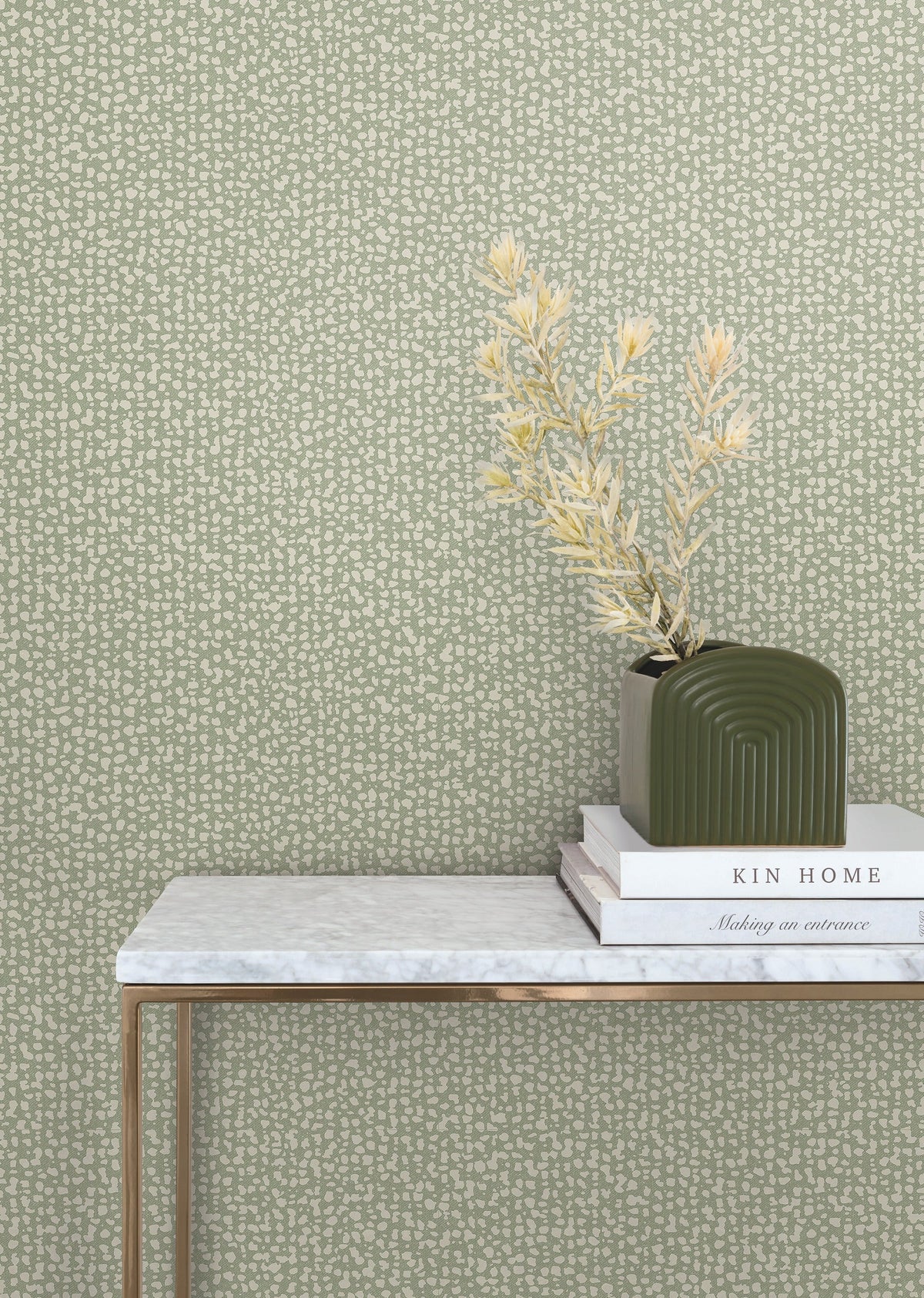 A marble-top table holds stacked books and a green vase with dried foliage, set against the Galaxies Wallpaper by York Wallcoverings in a light green, patterned design.