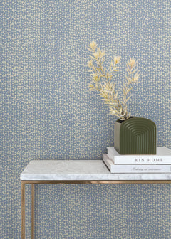 A green vase with dried yellow flowers sits on stacked books atop a marble console table with gold legs, set against York Wallcoverings’ Galaxies Wallpaper.