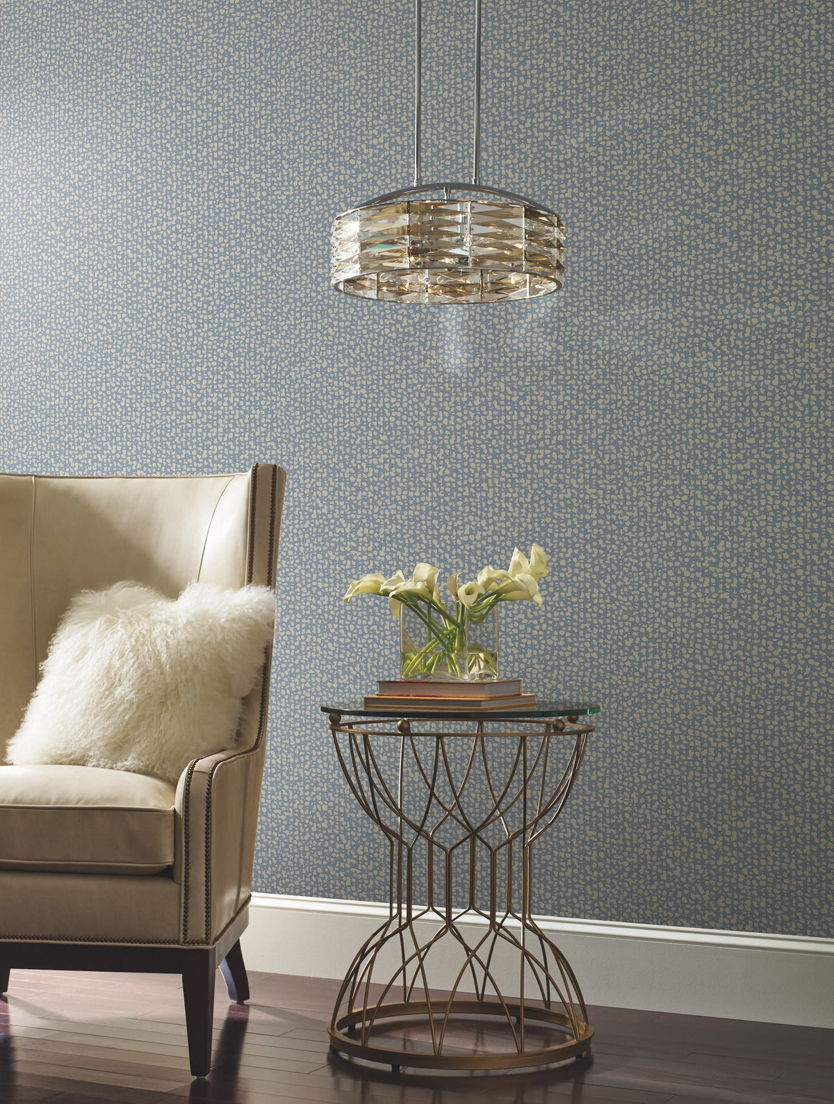 A beige armchair with a fluffy pillow sits next to a round metal side table holding yellow flowers and books, illuminated by a modern pendant light, all set against a blue Galaxies Wallpaper by York Wallcoverings.