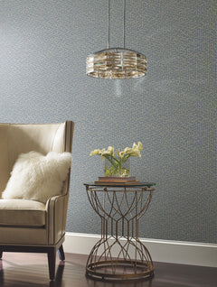 A beige armchair with a fluffy pillow sits next to a round metal side table holding yellow flowers and books, illuminated by a modern pendant light, all set against a blue Galaxies Wallpaper by York Wallcoverings.