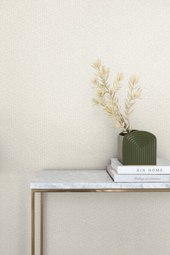 A console table with a marble top and gold legs stands against a wall covered in light, speckled Galaxies Wallpaper by York Wallcoverings, with a small vase of dried plants and two books on the surface.