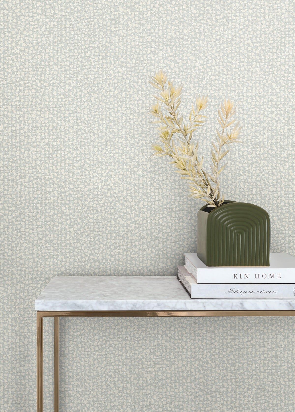 A green vase with dried foliage sits on stacked books atop a marble table with gold legs, set against a beige Galaxies Wallpaper by York Wallcoverings.