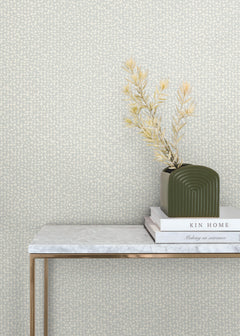 A green vase with dried foliage sits on stacked books atop a marble table with gold legs, set against a beige Galaxies Wallpaper by York Wallcoverings.