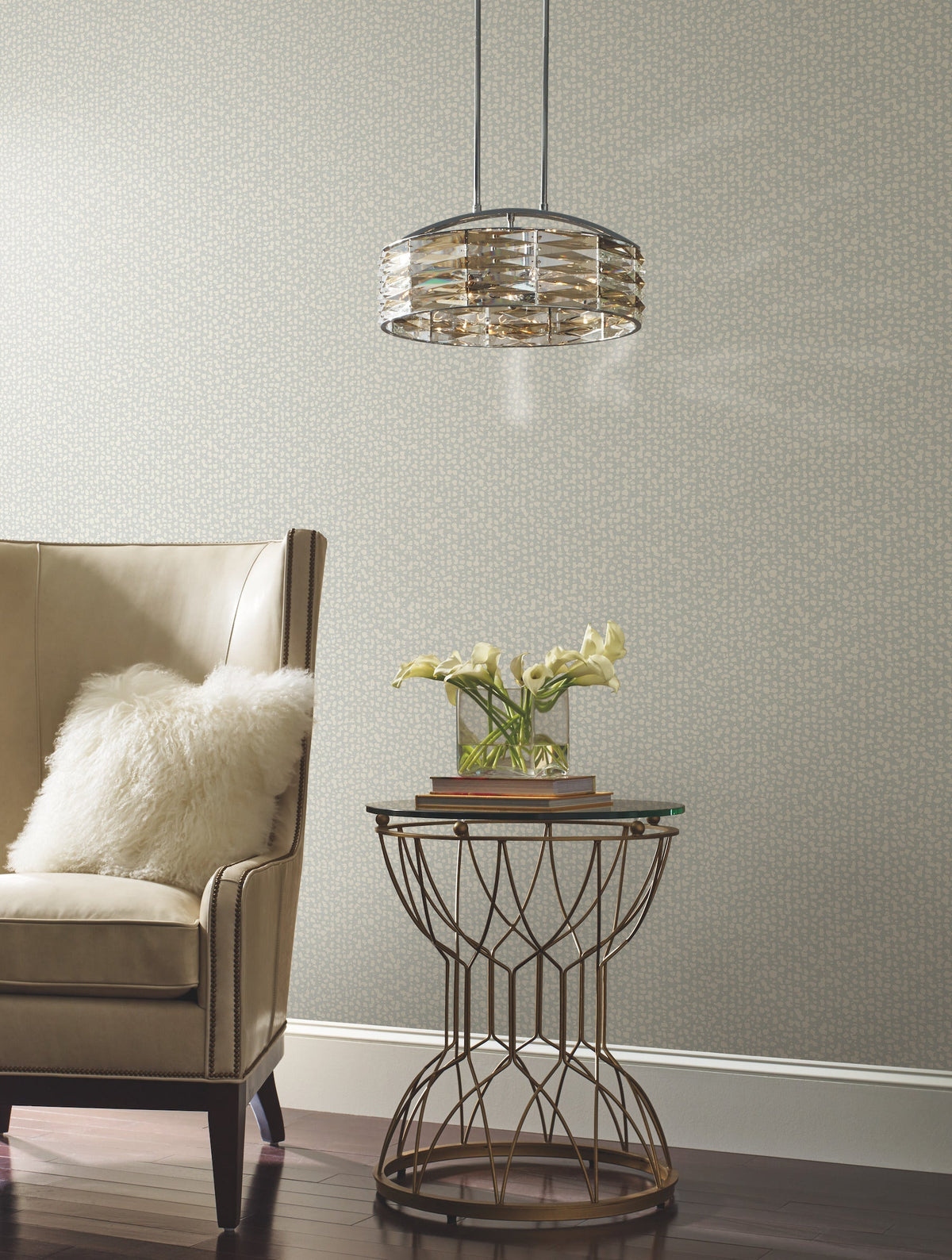 A beige armchair with a white pillow sits next to a round side table holding yellow flowers and books, beneath a modern pendant light, set against a wall adorned with York Wallcoverings’ Galaxies Wallpaper.