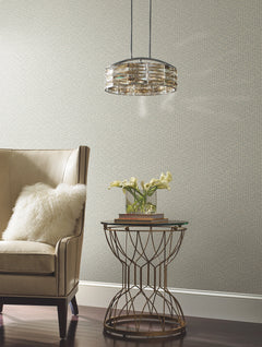 A beige armchair with a white pillow sits next to a round side table holding yellow flowers and books, beneath a modern pendant light, set against a wall adorned with York Wallcoverings’ Galaxies Wallpaper.
