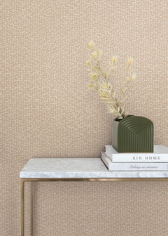 A green vase with dried stems sits on stacked books atop a slim, marble-topped table against a beige Galaxies Wallpaper by York Wallcoverings.