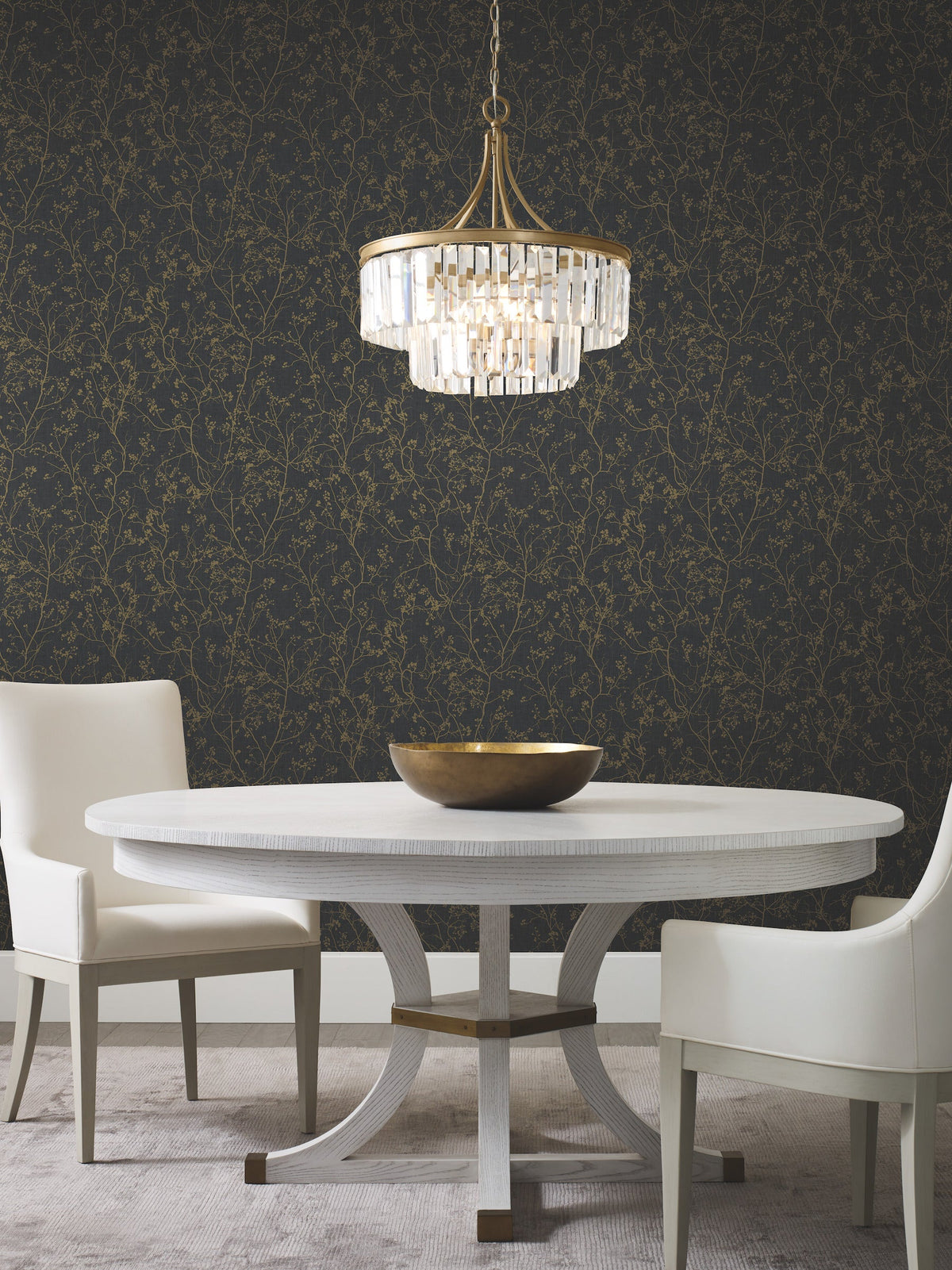 A modern dining area with a round white table, three white chairs, a gold bowl centerpiece, a crystal chandelier, and Luminous Branches Wallpaper by York Wallcoverings adorning the walls.