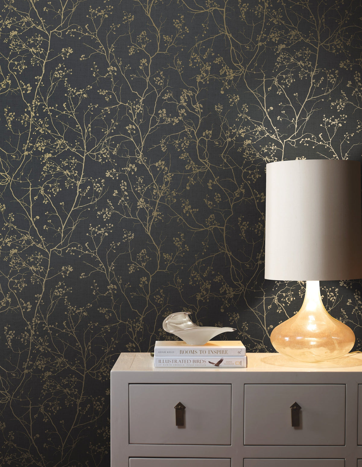 A beige lamp and a few books sit on a light-colored dresser in front of York Wallcoverings’ Luminous Branches Wallpaper, featuring a delicate gold botanical pattern.