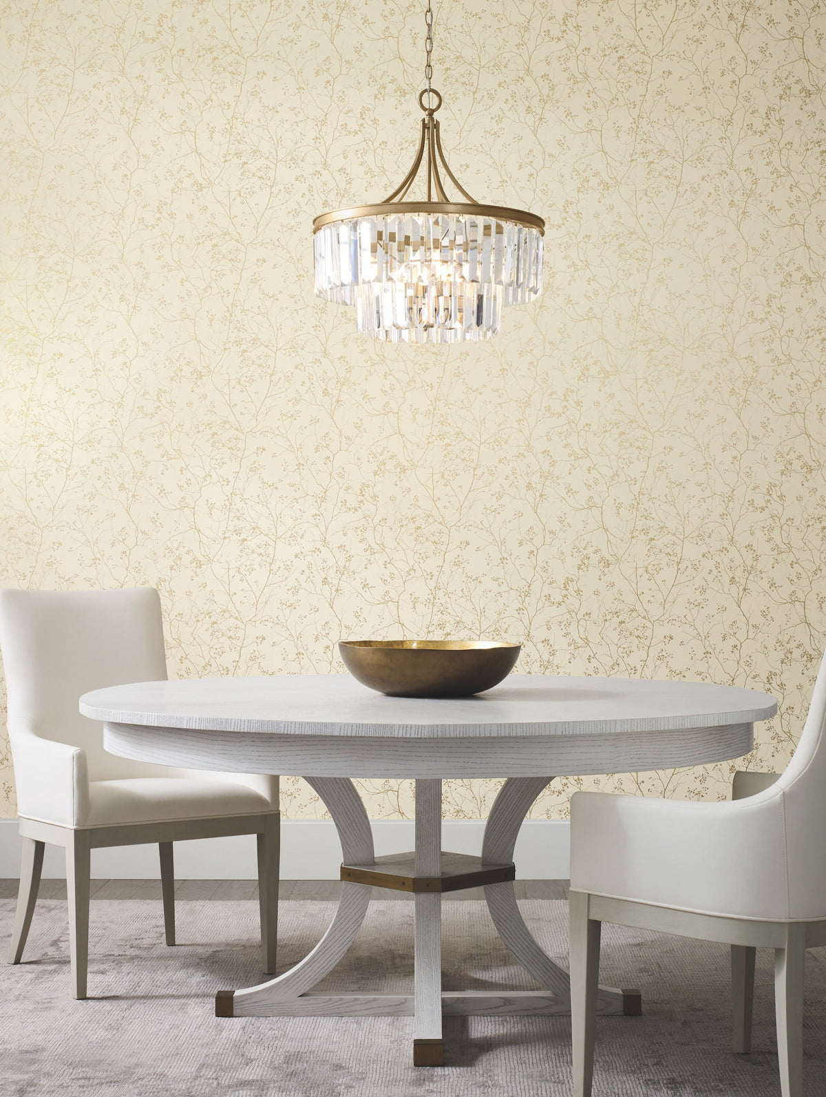 A round white table with a bronze bowl sits between two white chairs under a crystal chandelier, set against Luminous Branches Wallpaper by York Wallcoverings.