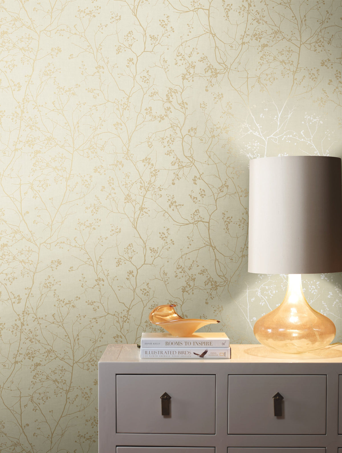 A glass lamp with a white shade sits on a beige dresser next to a book and glasses, set against York Wallcoverings’ Luminous Branches Wallpaper featuring a delicate gold floral pattern.