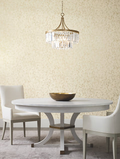 A round white table with a gold bowl sits between two white chairs. Above, a gold and crystal chandelier hangs, while the background showcases York Wallcoverings’ Luminous Branches Wallpaper.