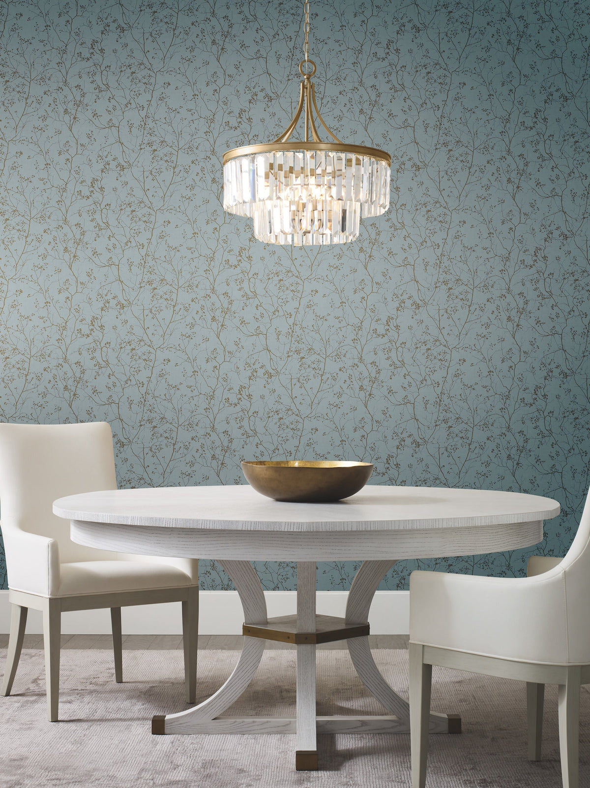 Elegant dining area with a round white table, two white chairs, a gold decorative bowl, and a crystal chandelier. The background features York Wallcoverings’ Luminous Branches Wallpaper in blue with a delicate gold floral pattern.