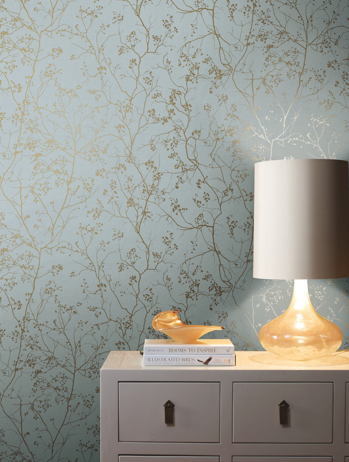 A beige lamp rests on a light gray dresser adorned with books and a decorative dish, set against York Wallcoverings’ Luminous Branches Wallpaper in blue, which features delicate gold branches and small blossoms.