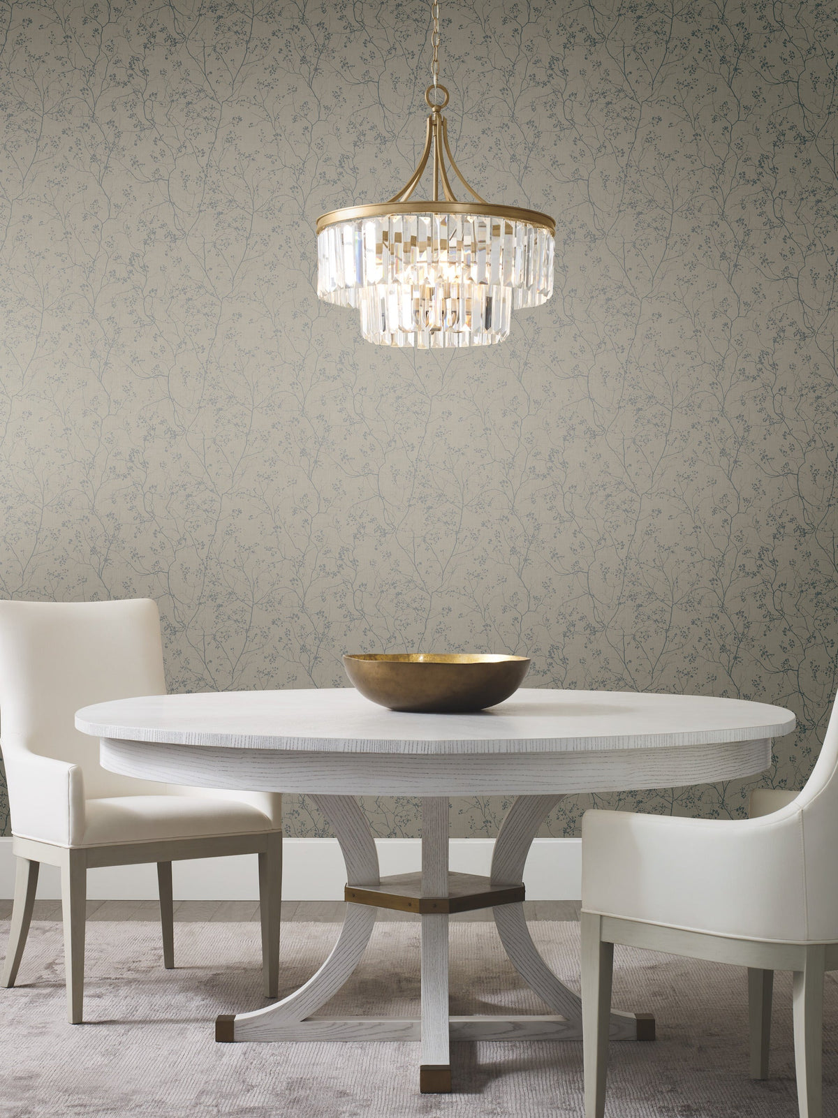 A round white dining table with two white chairs, a gold bowl centerpiece, a crystal chandelier overhead, and York Wallcoverings’ Luminous Branches Wallpaper in gray as a subtle floral-patterned backdrop.