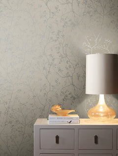 A beige dresser adorned with books, a glass bird, and a lit lamp sits against walls covered in York Wallcoverings’ Luminous Branches Wallpaper.