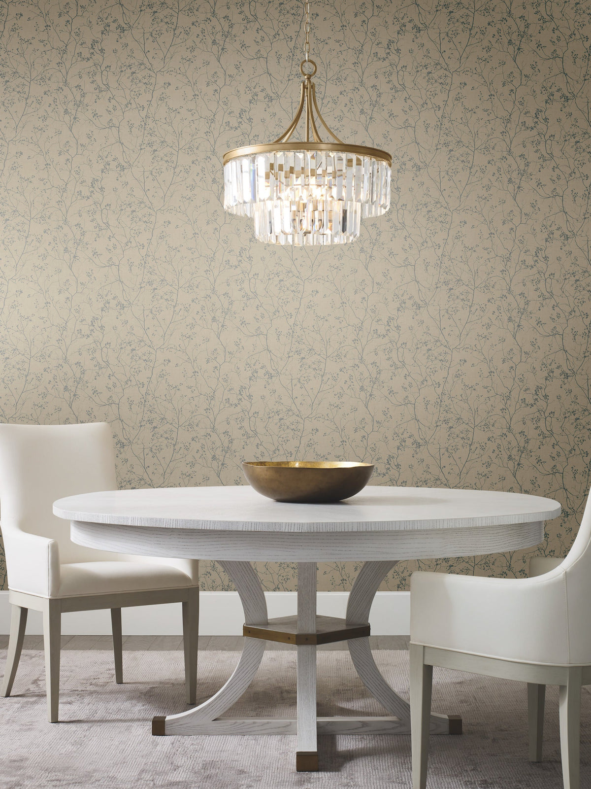 A round white table with two white chairs, a gold bowl centerpiece, and a crystal chandelier above, set against Luminous Branches Wallpaper by York Wallcoverings.