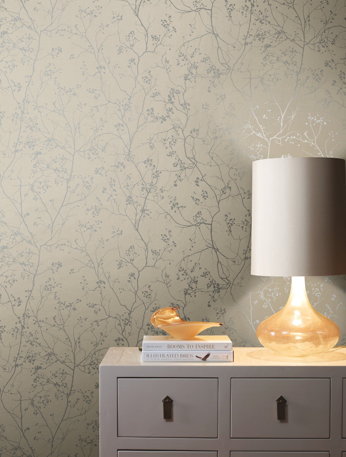 A lit glass lamp, books, and a small decorative object sit on a light-colored dresser against beige York Wallcoverings Luminous Branches Wallpaper featuring a delicate branch pattern.