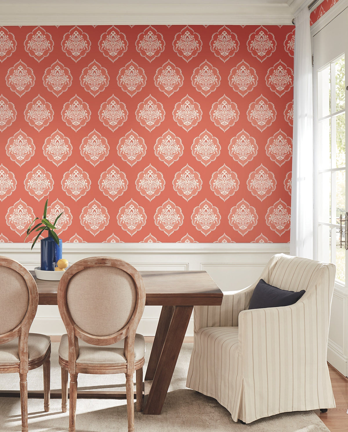Dining room with a wooden table, four upholstered chairs, a blue vase with flowers, and York Wallcoverings’ Signet Medallion Damask Wallpaper in red and white. Large windows on the right let in natural light.
