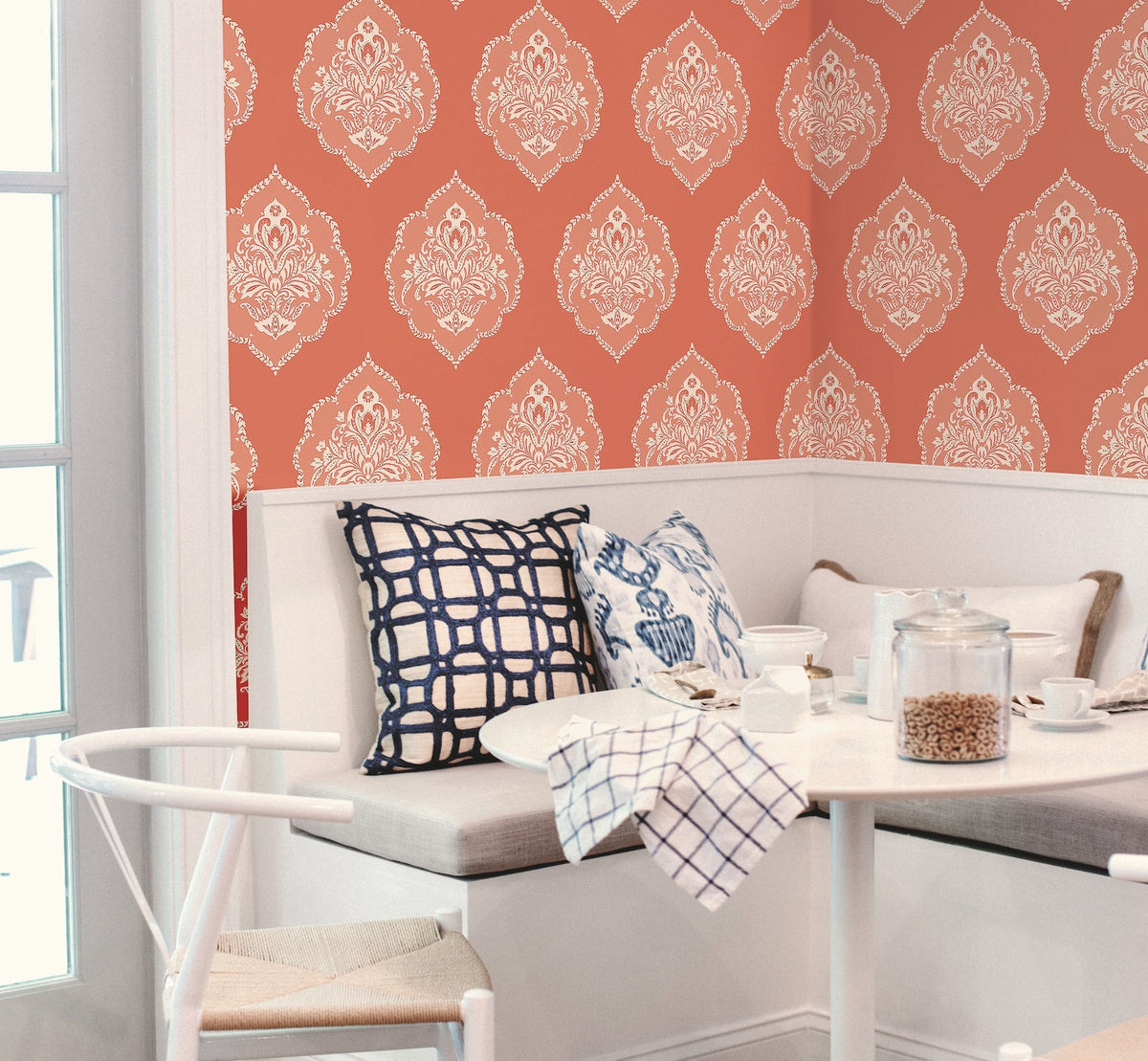 A cozy breakfast nook featuring a white bench, blue-patterned pillows, and a small table set for breakfast, with York Wallcoverings’ Signet Medallion Damask Wallpaper in a rich red pattern providing an elegant backdrop. Light streams in through a glass door on the left.