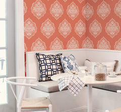 A cozy breakfast nook featuring a white bench, blue-patterned pillows, and a small table set for breakfast, with York Wallcoverings’ Signet Medallion Damask Wallpaper in a rich red pattern providing an elegant backdrop. Light streams in through a glass door on the left.