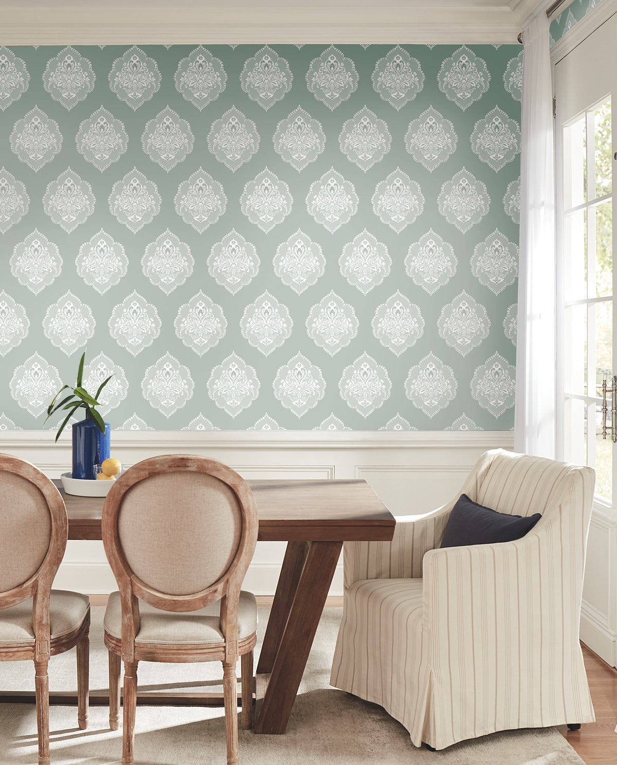 A dining room with a wooden table, three upholstered chairs, one striped armchair, and a blue vase with greenery. The wall features York Wallcoverings’ Signet Medallion Damask Wallpaper in blue and white. Sunlight streams in from large windows.