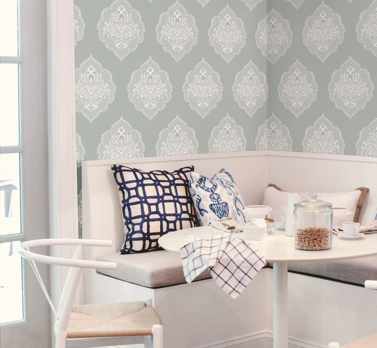 A cozy breakfast nook features a white table, patterned pillows, and York Wallcoverings’ Signet Medallion Damask Wallpaper in light gray and white. By the window, jars, bowls, and a tea towel rest on the tabletop.
