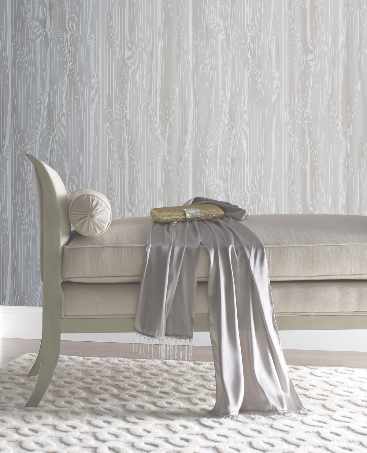 An upholstered bench with a small bolster pillow, a gold clutch, and a draped gray scarf, set against York Wallcoverings Candice Olson Fantasy Faux Bois Wallpaper featuring a white wood-grain pattern.
