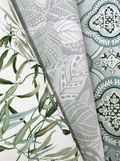 Three overlapping rolls of York Wallcoverings’ Willow Grove Wallpaper: one featuring green leaves, another with a gray and white abstract leafy pattern, and a third showcasing a blue and white ornate geometric design with floral motifs.