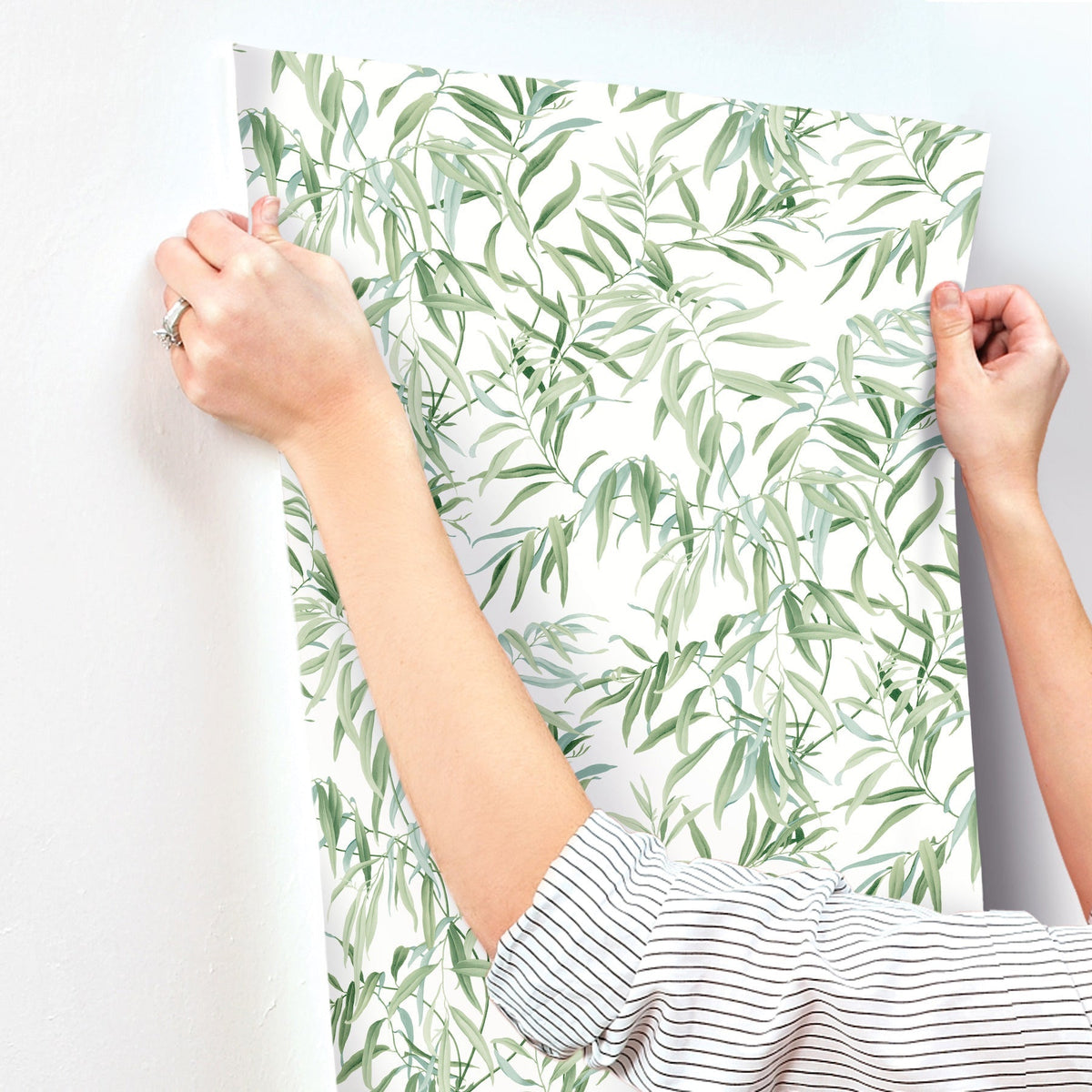 A person applies York Wallcoverings’ Willow Grove Wallpaper, featuring a green leafy vine pattern, to a white wall. Only their hands and arms—clad in a striped shirt with a ring—are visible.