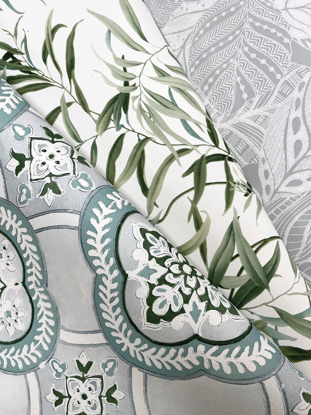 Three different patterned wallpapers overlap: York Wallcoverings’ Willow Grove Wallpaper featuring green leafy branches, one with light gray intricate abstract designs, and another with teal and white ornate floral motifs set against a gray background.