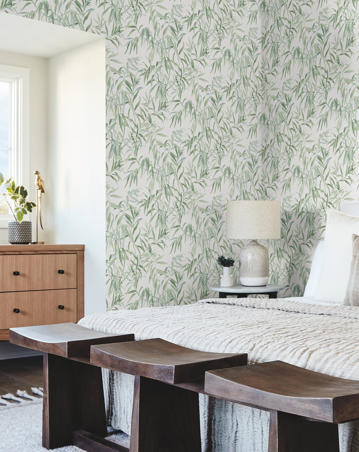A bedroom features Willow Grove Wallpaper by York Wallcoverings, a wooden bench, and a dresser. White bedding, a textured blanket, a lamp, and sunlit windows complete the cozy, modern decor.