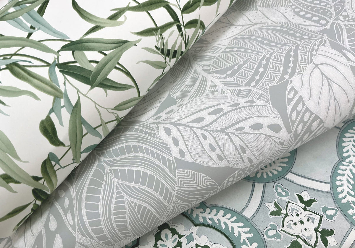 Three rolls of Willow Grove Wallpaper by York Wallcoverings in assorted botanical designs: one featuring green leaves, one with white outlined leaves on a gray background, and one adorned with intricate floral patterns in green, blue, and white.