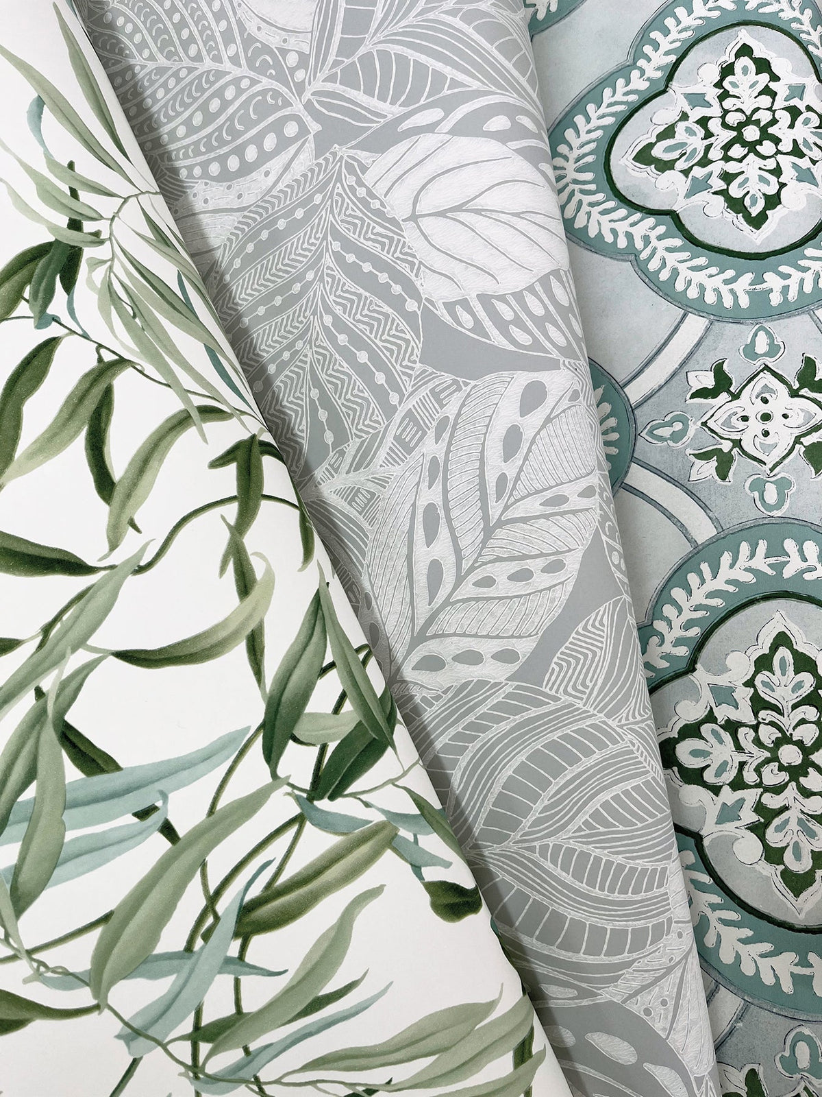 Three rolled wallpapers from York Wallcoverings: the Willow Grove Wallpaper featuring green leafy vines, a design with intricate gray and white leaves, and a pattern showcasing blue and white floral damask on a light blue background.