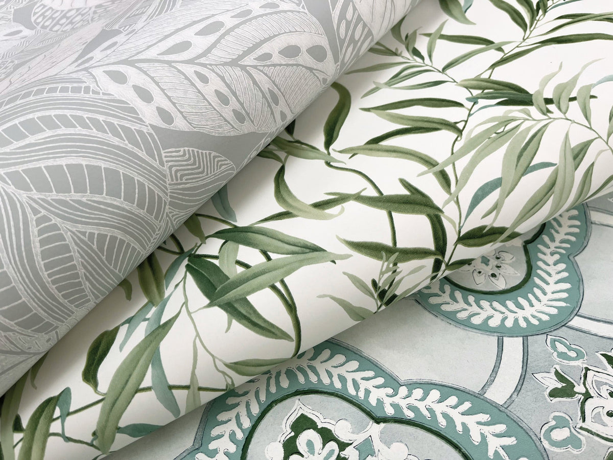 Three sheets of decorative wallpaper overlap: one features gray leaf designs, one showcases green leafy branches on white, and one displays the “Willow Grove Wallpaper” by York Wallcoverings with its blue and white intricate pattern.