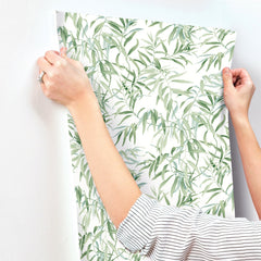 A person wearing a striped shirt hangs Willow Grove Wallpaper by York Wallcoverings, featuring a leafy green pattern, on a white wall.