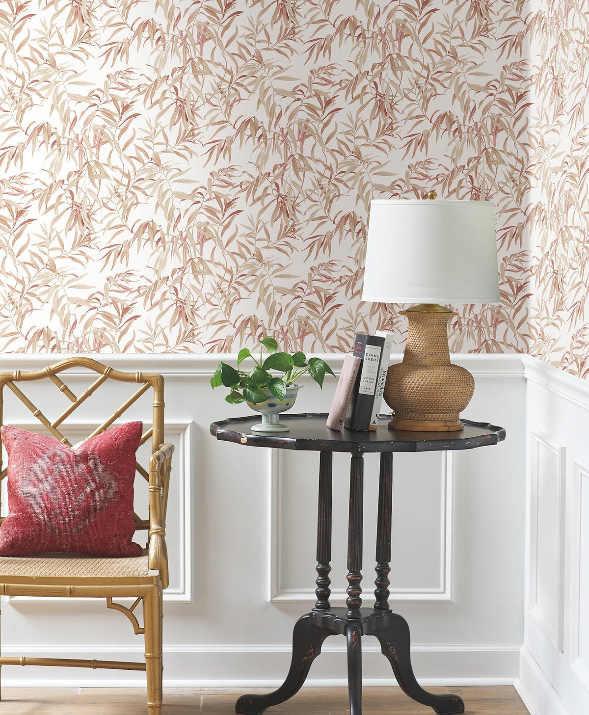 A bamboo chair with a red pillow sits next to a round black table holding a plant, lamp, and books. The background features Willow Grove Wallpaper by York Wallcoverings and white wainscoting.