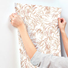 A person in a striped shirt applies York Wallcoverings’ Willow Grove Wallpaper, featuring a beige and light brown leafy pattern, to a white wall.